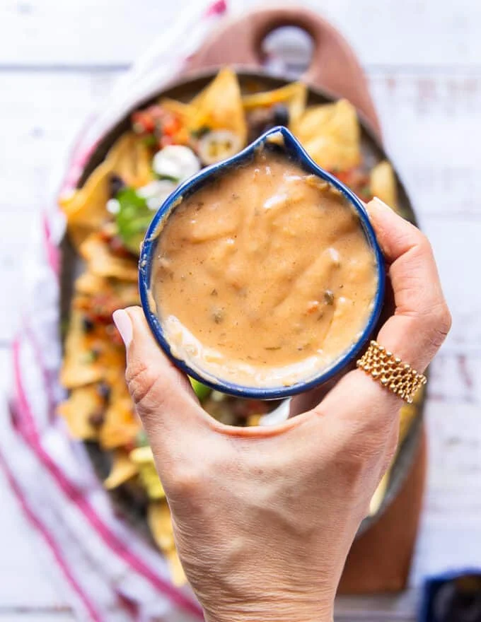 A hand holding a bowl of nacho cheese sauce ready to drizzle it over the loaded ground beef nachos