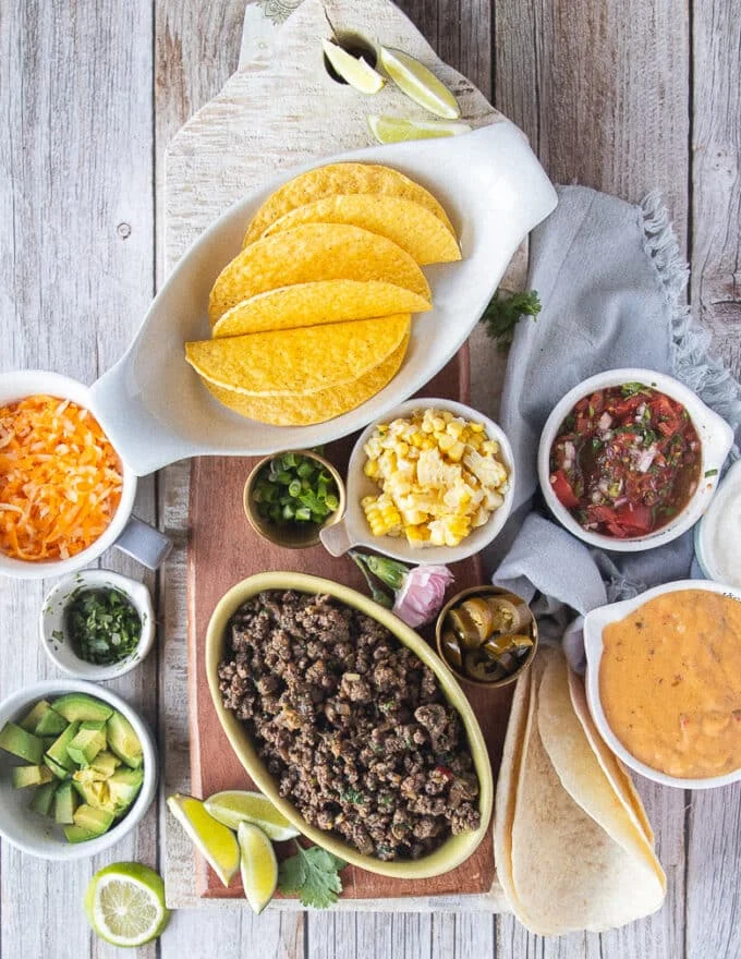 ingredients for ground beef tacos on a board including a bowl of ground beef, some taco shells, some diced avocados, some cheese sauce, some salsa, some cilantro, some green onions and a bowl of cheese, a bowl of jalapeno