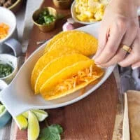 A hand holding a hard taco shell in a baking sheet and adding cheese to the taco shell to start assembling the ground beef tacos