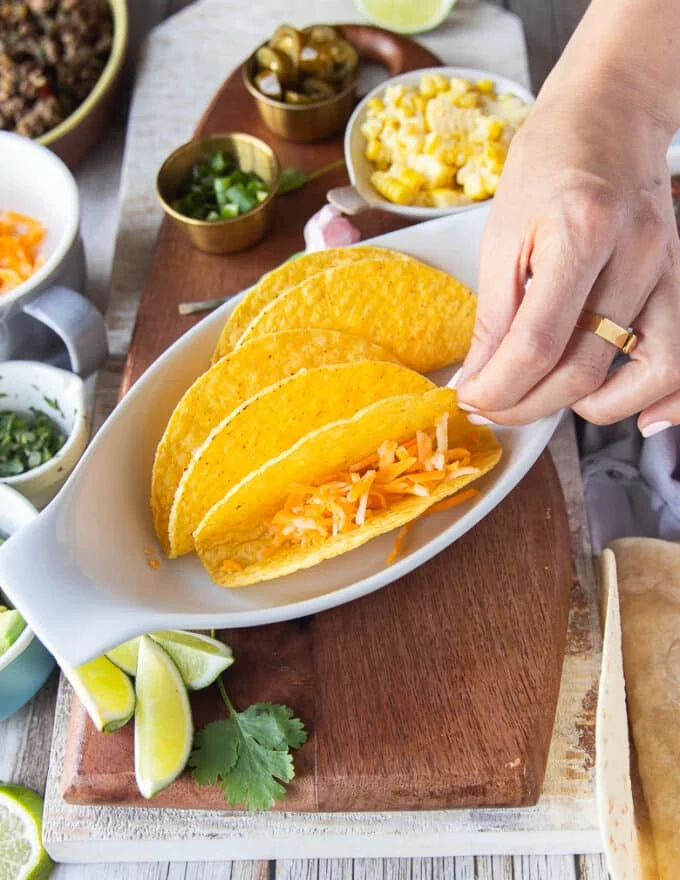 A hand holding a hard taco shell in a baking sheet and adding cheese to the taco shell to start assembling the ground beef tacos