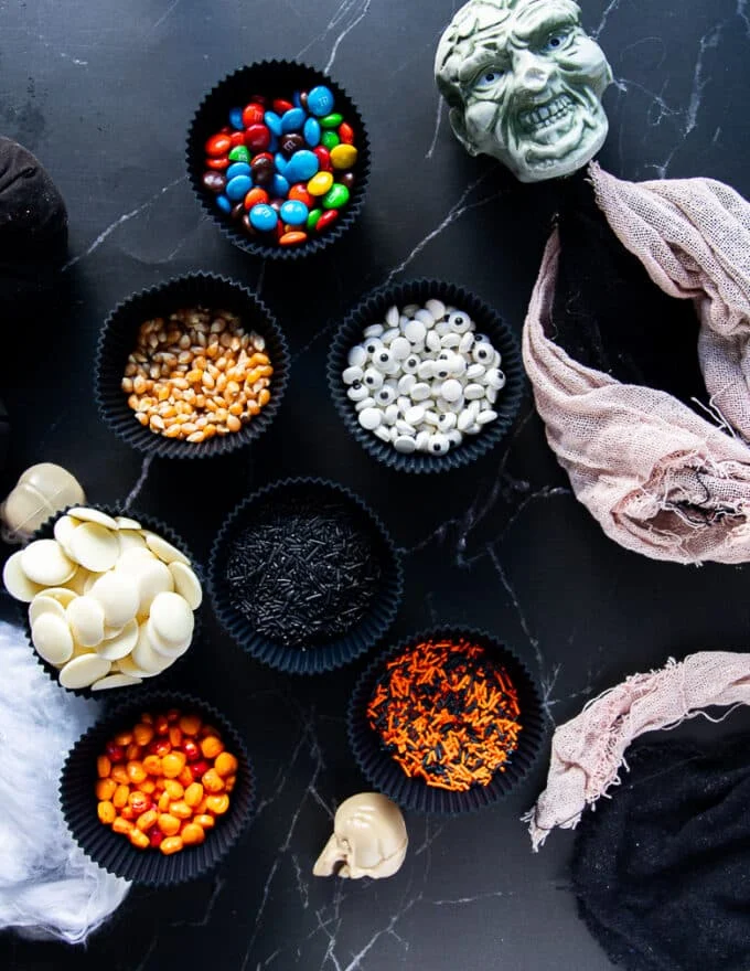 ingredients for the halloween popcorn including bowl of raw corn kernels, white chocolate wafers, bowls of sprinkles, bowls of candies, bowls of colored chocolate