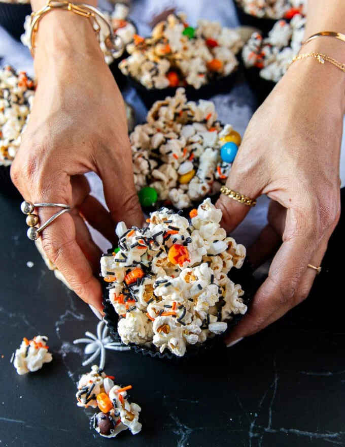 A hand holding a cup of halloween popcorn with some falling around it and a mini plastic skull surrounding the hand for a spooky feel