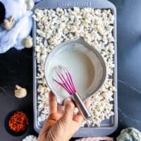 A hand holding a bowl of melted white chocolate to pour over the popcorn on a baking sheet