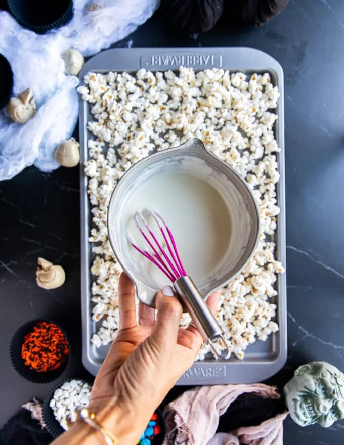 A hand holding a bowl of melted white chocolate to pour over the popcorn on a baking sheet