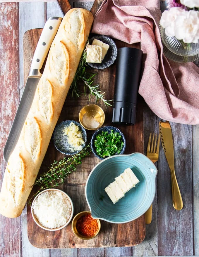 Ingredients for air fryer garlic bread on a borad including a loaf of baguette bread, some butter in a bowl, some fresh herbs, some grated parmesan cheese, some salt, pepper and fresh garlic minced , a bowl of garlic powder and tea towel