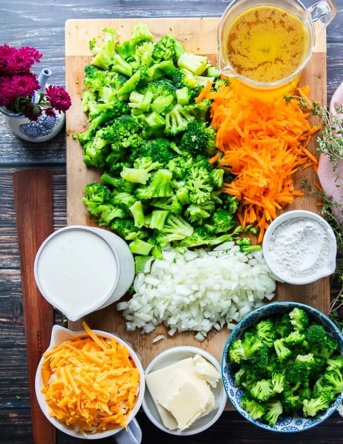 ingredients for broccoli cheddar soup recipe including broccoli florets on a bowl, some shredded cheese, a bowl or milk, a bowl of cream, a bowl of flour, diced onions in a board, minced some butter on a plate, garlic in a bowl, a jar of stock , seasoning in a bowl, some mustard in a bowl