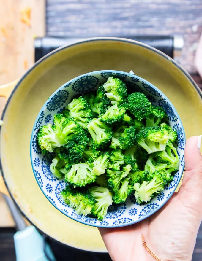 A hand holding a bowl of mini broccoli florets chopped finely to add to the final pot