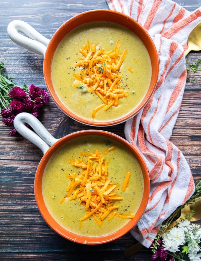 two bowls of broccoli cheddar soup on a board with a tea towel in the middle
