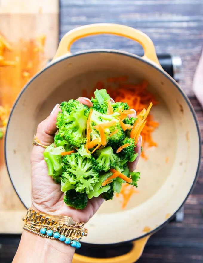 A hand holding in some broccoli florets and adding them to the base of the soup pot