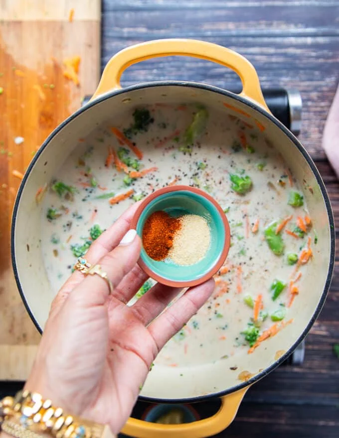 A hand holding some seasoning like garlic powder, paprika in a bowl and seasoning the soup