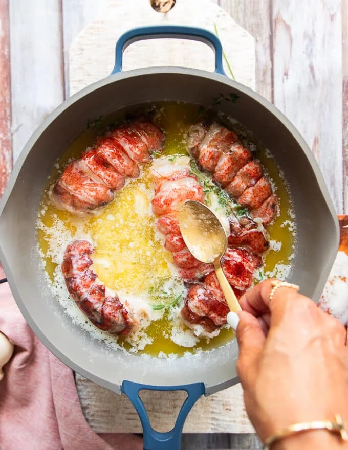 A spoon drenching the tops of the lobster tails with the butter sauce to help it cook the way through