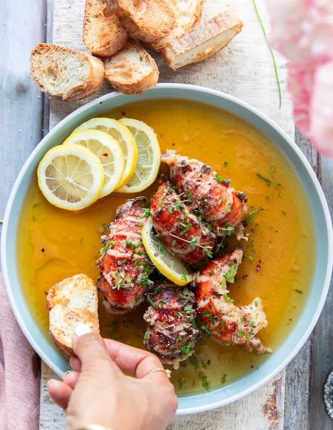 A hand holding a piece of garlic bread and dipping it into the plate of butter poached lobster to mop up the sauce