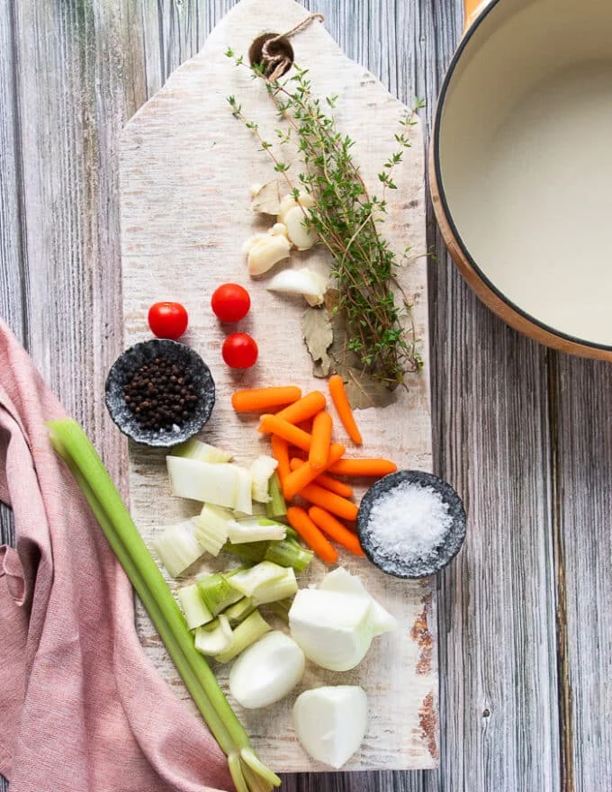 stock ingredients on a white board including some carrots, celery, onions, garlic, bay leaves, fresh herbs, baby tomatoes, coarse salt and pepper