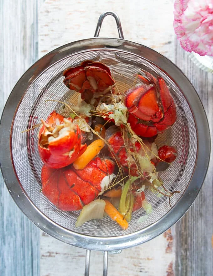The lobster stock is passed through a strainer showing the lobster shells, all the veggies and herbs in the strainer, and a clear golden liquid left