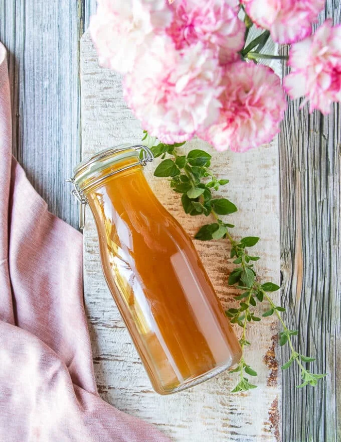 A sealed glass bottle of lobster stock on a white board surrounded by herbs and flowers