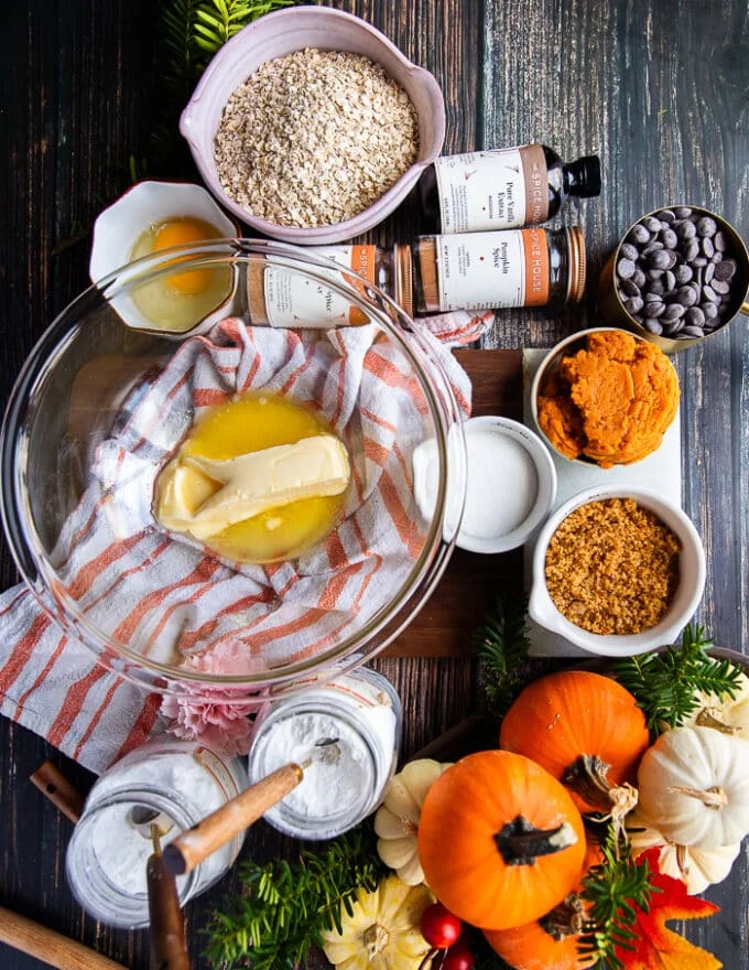 ingredients for pumpkin oatmeal cookies in a board including soft butter in a bowl, some white sugar, brown sugar in a bowl, vanilla, pumpkin spice, pumpkin puree, baking powder, baking soda, flour, oats in a bowl and one egg yolk