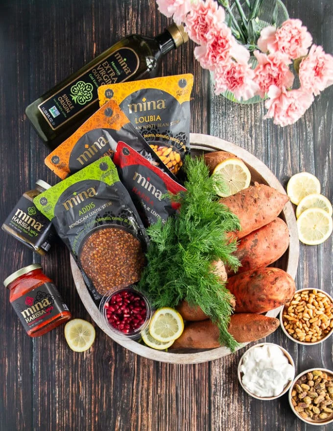 ingredients for stuffed sweet potatoes including sweet potatoes in a basket, a bunch of dill, a bag of lentil, some harissa or hot sauce, salt and pepper, some feta in a bowl, some pine nuts in a bowl, some olive oil and pomegranate