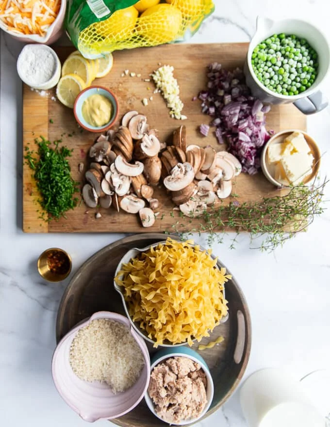 Ingredients on a large marble for tuna casserole. Caned tuna in a bowl, cheese in a bowl, some milk, some butter in separate bowls, the egg noddles uncooked in a bowl, sliced mushrooms on a board with diced onions, some garlic minced, a bowl of dijon mustard, some frozen peas in a bowl,some lemon slices, a bowl of seasoned panko, some half n half in a cup