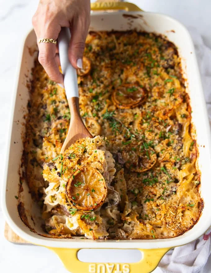 A hand using a large spoon to scoop up some tuna casserole showing the texture of the noodles and tuna and the crunchy topping