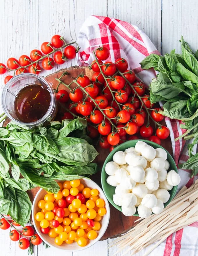 Ingredients for Caprese Wreath including a large bowl of baby mozzarella, a bowl of cherry tomatoes, a bowl of orange colored tomatoes, and three large bunches of fresh basil