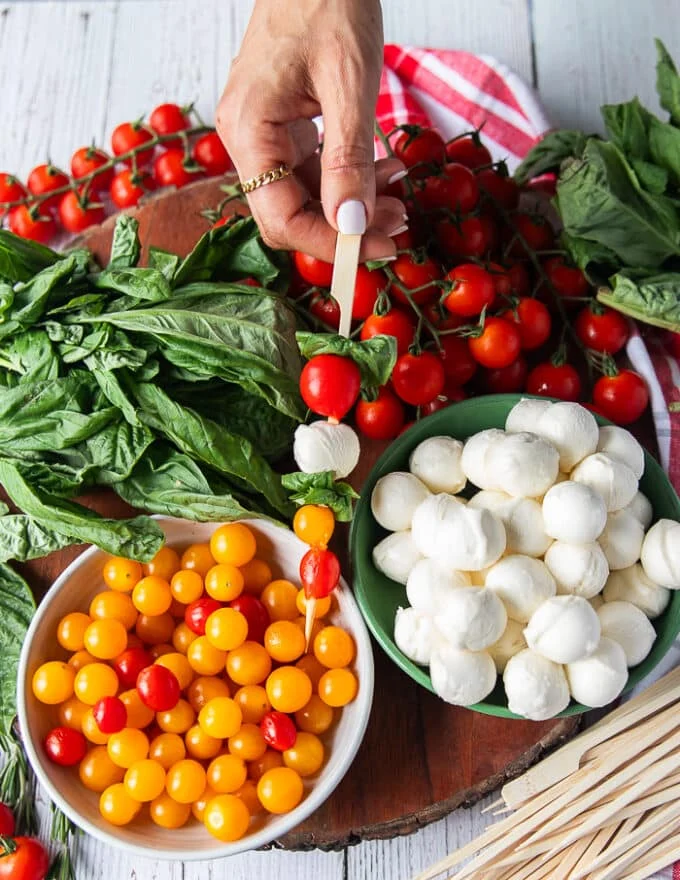 A hand holding a bamboo skewer with a basil leaf skewered followed by cherry tomato, bocconcini , more basil, baby orange and yellow tomatoes and another basil leaf