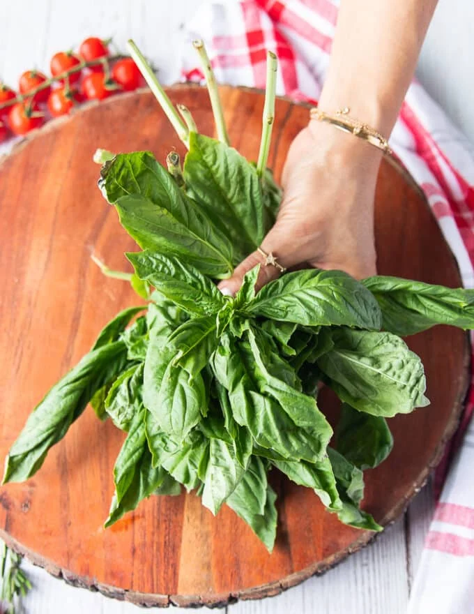 A hand holding three bunches of fresh basil leaves on a round wooden board to make the base of the caprese wreath