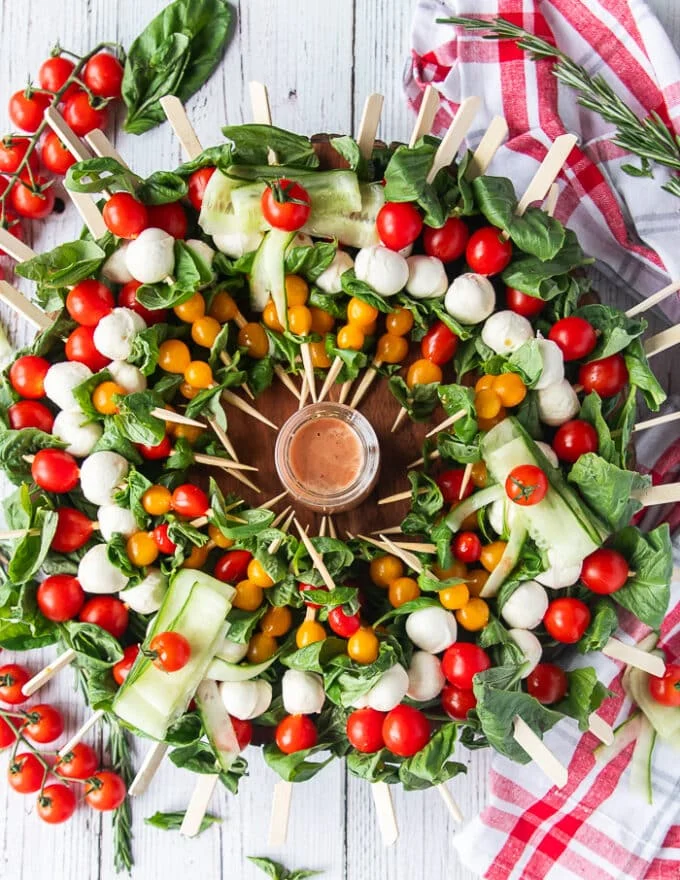 A whole caprese wreath with skewers of tomatoes, basil and mini mozzarella cheese on a bed of basil leaveas and a small bowl of vinaigrette in he middle