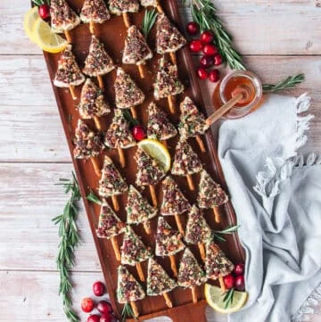 A board with small cheese Christmas trees coated in pecan, herb cranberry and lemon zest and each has a pretzel stick as a base. The board is surrounded by fresh cranberries, rosemary, lemon slices and a tea towel and looks very festive