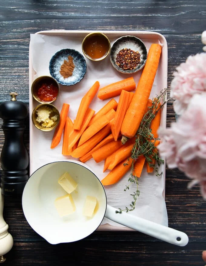 Ingredients for honey glazed carrots including carrots, a bowl with butter, a bowl with honey, some seasoning, minced garlic in a bowl, some fresh herbs, a bowl with chilli flakes, a bowl with harissa and some cinnamon in a bowl