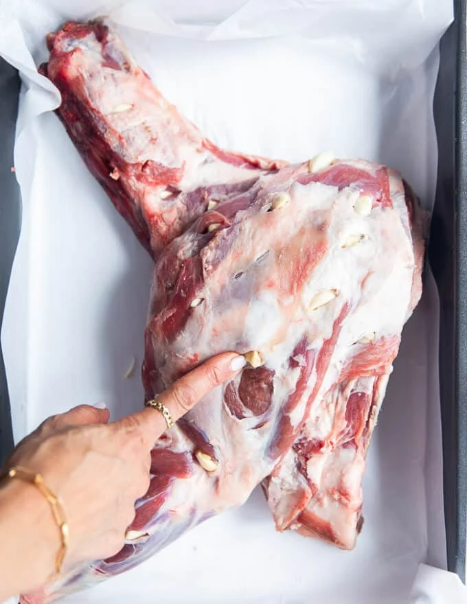 A hand inserting whole garlic cloves into the slits along the lamb shoulder