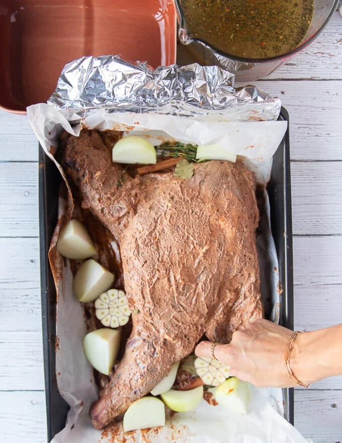 Onions and whole garlic head added around the lamb along with the whole spices in the roasting pan with the marinated lamb shoulder in the middle