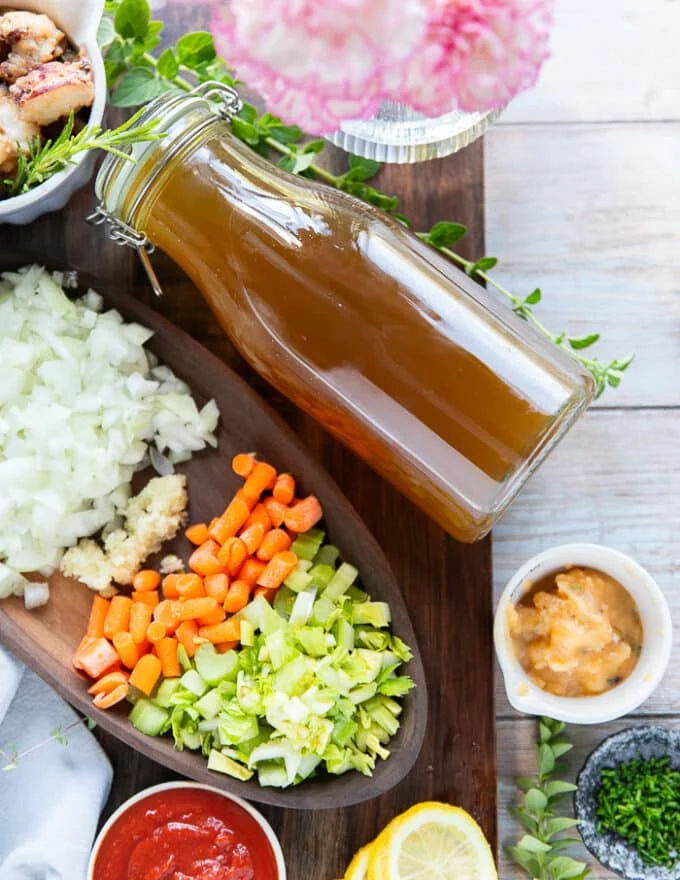 Ingredients to make lobster bisque on a wooden board. including a jar of lobster stock, some diced onions in a bowl, diced celery, diced carrot, minced garlic, lobster butter, cooked lobster meat, some cream and fresh herbs