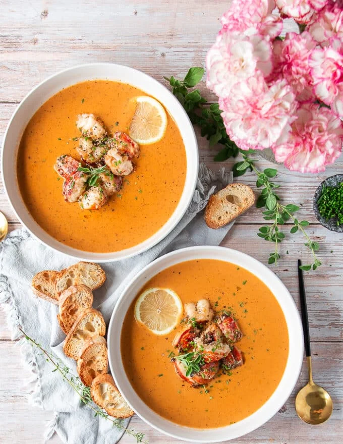 two bowls of lobster bisque plated on a board with some garlic bread on the side, some lemon wedges, a small pot of flowers and some fresh herbs around it