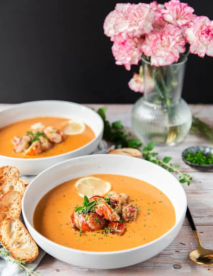 Side view of tow lobster bisque bowls surrounded by garlic bread and topped with lots of succulent lobster and a lemon slice
