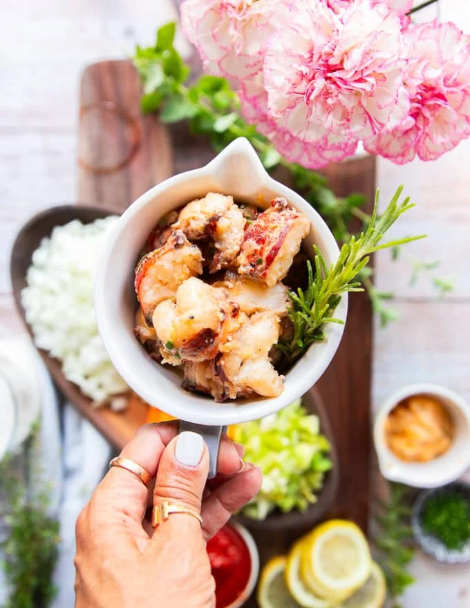 A hand holding a bowl of cooked lobster in butter to show how perfectly cooked and succulent it looks on the board of recipe ingredients