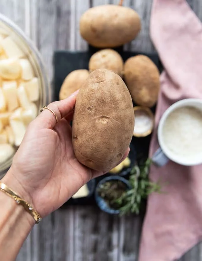A hand holding a potato over a surface with some cream, milk, butter, salt and pepper , herbs and garlic
