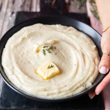 A hand holding a plate of mashed potatoes close up showing a dab of butter melting on the top and a rich creamy spread up mashed potatoes on a plate