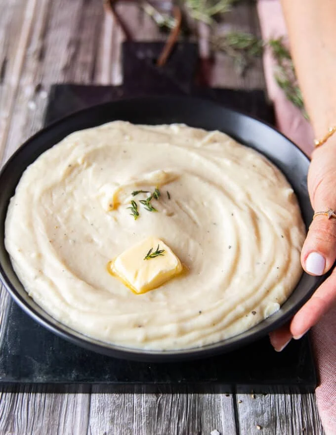 A hand holding a plate of mashed potatoes close up showing a dab of butter melting on the top and a rich creamy spread up mashed potatoes on a plate