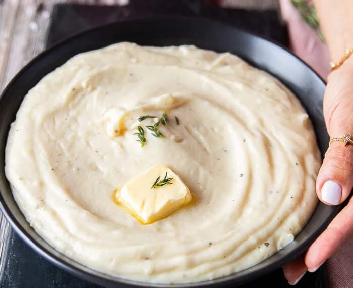 A hand holding a plate of mashed potatoes close up showing a dab of butter melting on the top and a rich creamy spread up mashed potatoes on a plate