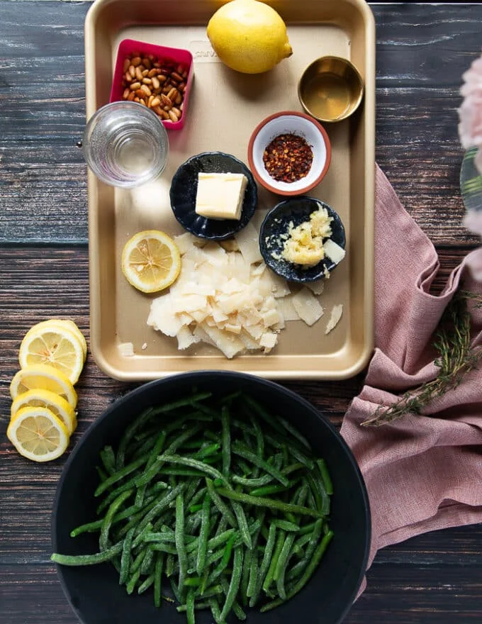 Ingredients to make sautéed green beans including a bowl of green beans, a tray with some bowls of parmesan, garlic, butter, seasoning, toasted pine nuts. herbs and lemon zest