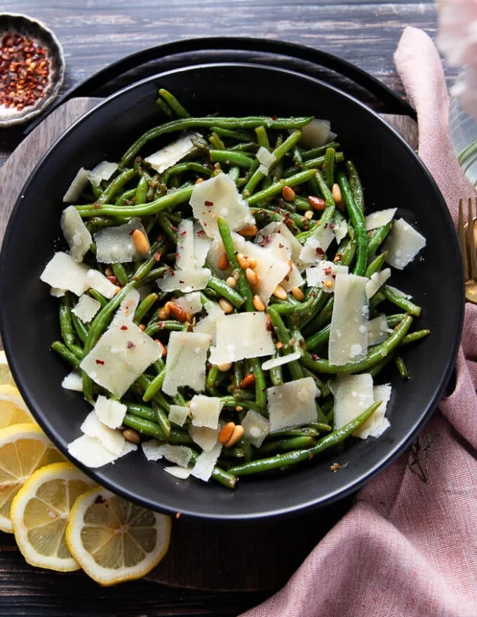 close up of a plate of perfectly sautéed green beans garnished with parmesan shavings, some lemon zest, chilli flakes and toasted nuts