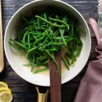 The green beans are added to the sauce pan over the butter and garlic and a wooden spoon is mixing them