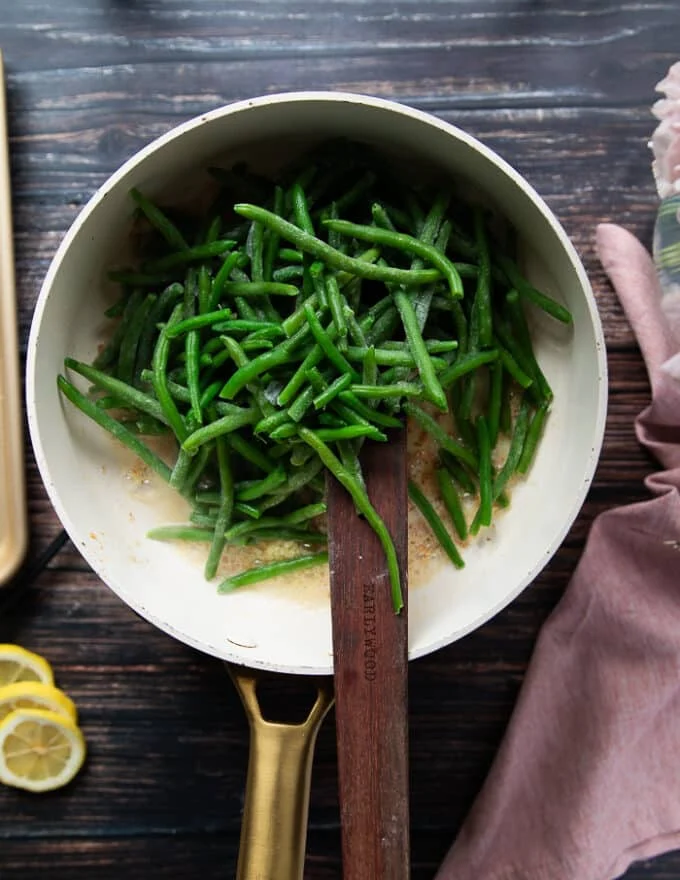 The green beans are added to the sauce pan over the butter and garlic and a wooden spoon is mixing them