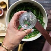 A hand pouring in such a few tablespoons of water over the green beans