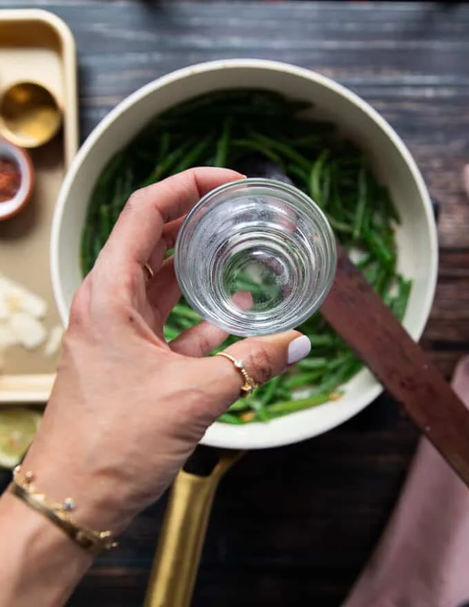 A hand pouring in such a few tablespoons of water over the green beans