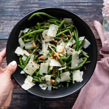 A hand holding a plate of perfect sautรฉed green beans. Showing the crisp tender green beans on a plate with shaved parmesan cheese, some nuts, bits of garlic and lemon zest