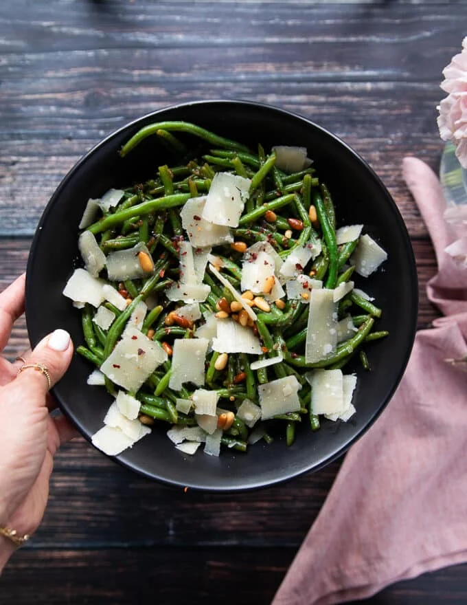 A hand holding a plate of perfect sautéed green beans. Showing the crisp tender green beans on a plate with shaved parmesan cheese, some nuts, bits of garlic and lemon zest