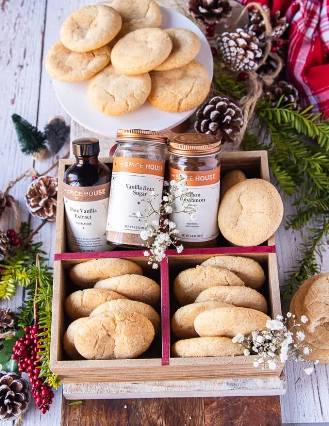 A large cookie box filled with sugar cookies and some jars of vanilla extract and vanilla bean sugar surrounded by pine cones, some flowers and a plate of more sugar cookies