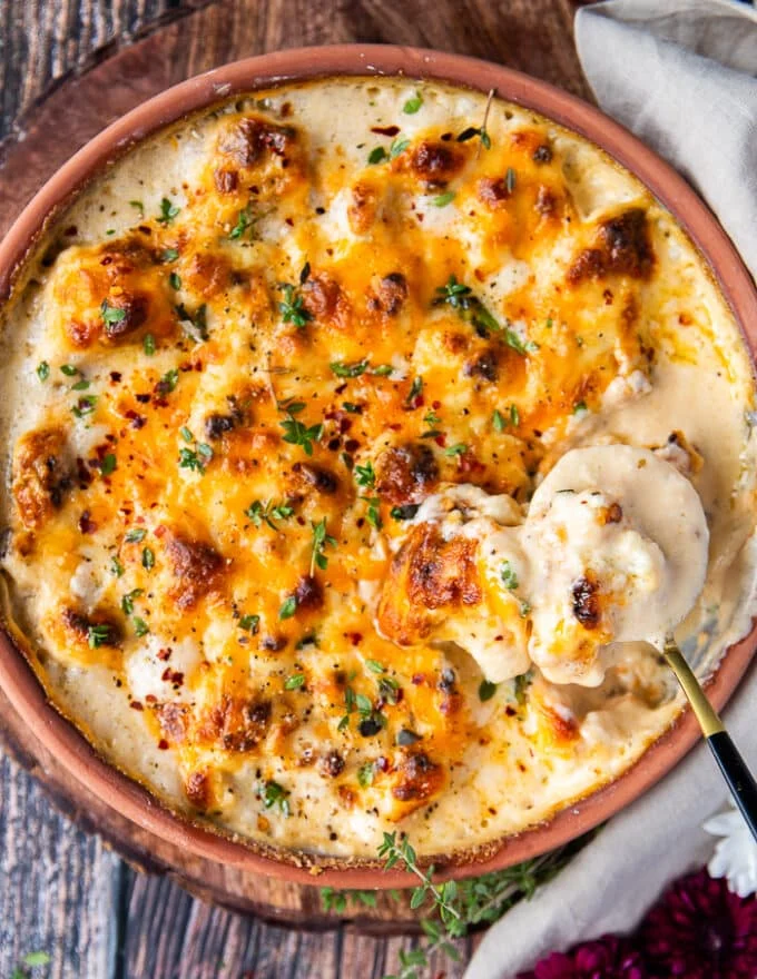 A spoon showing off the creamy cauliflower casserole showing the florets, and the creamy cheese sauce around it in a casserole dish