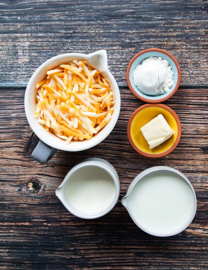 bechamel sauce ingredients on a wooden board including cups of milk, a cup of cream, a bowl of cheese, a small bowl of butter and small bowl of flour